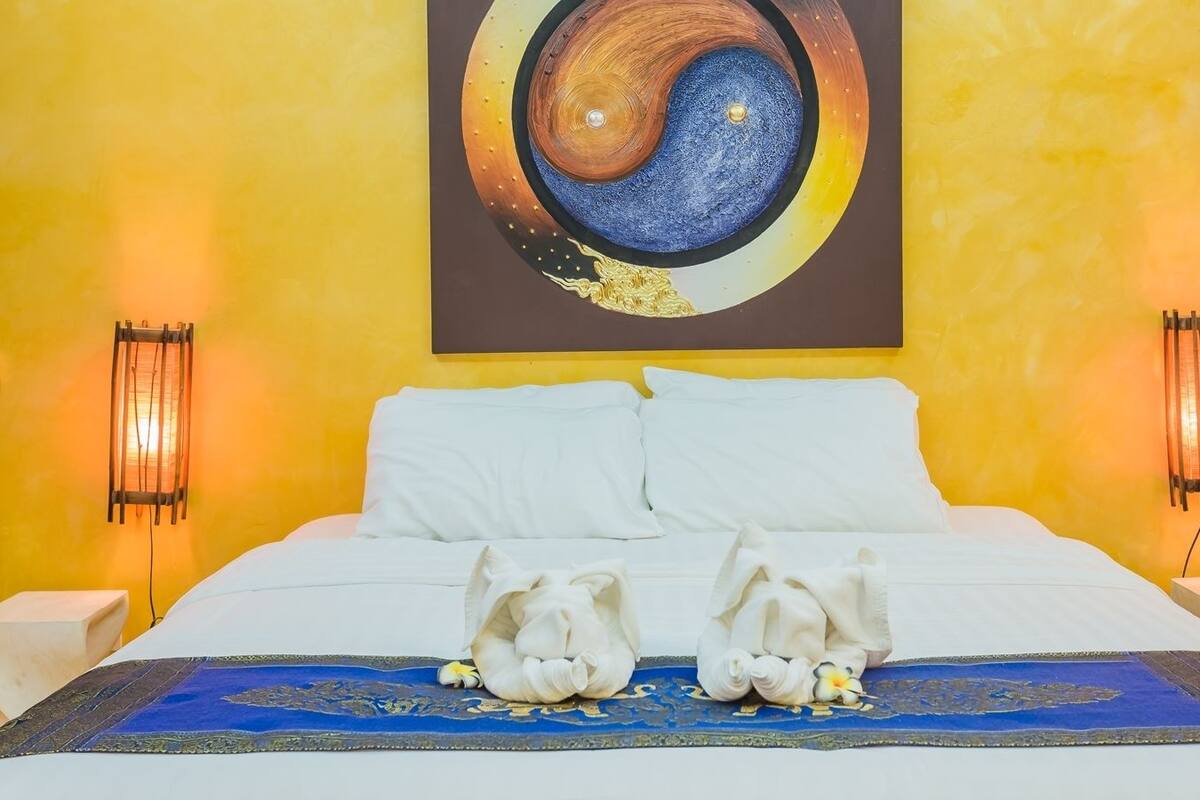 Une chambre d’hôtel avec un lit, des oreillers blancs, une couvre-lit aux motifs bleus et dorés, et un grand tableau représentant le symbole du yin et du yang au mur.