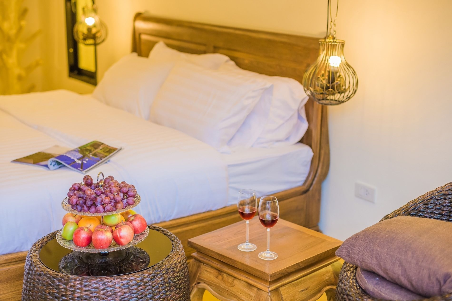A neatly made bed with white linens, a fruit display on a round table, and two glasses of red wine on a wooden tray.