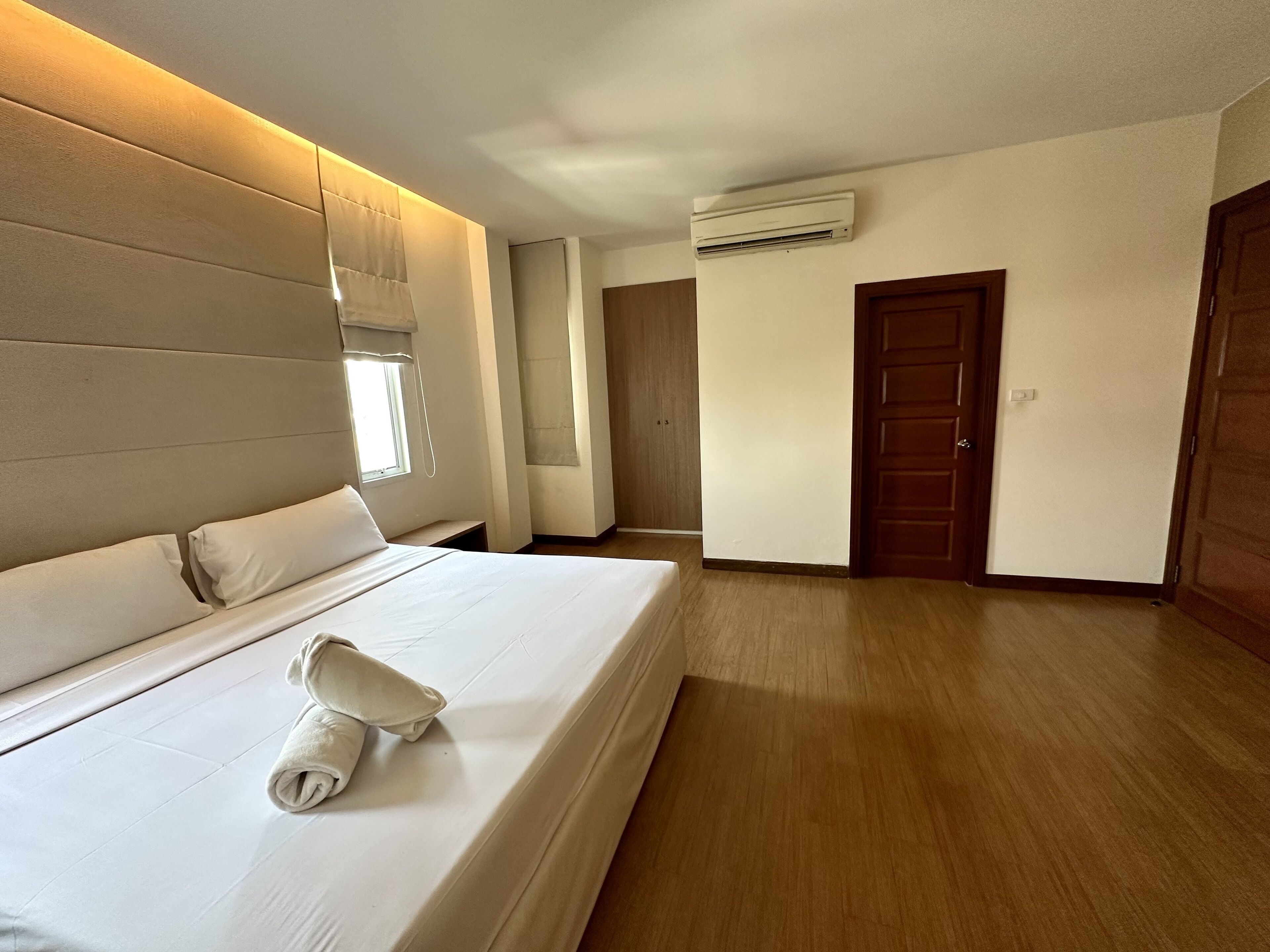 A hotel room with a large bed, a bedside table, a window with blinds, an air conditioning unit, and a wooden door.