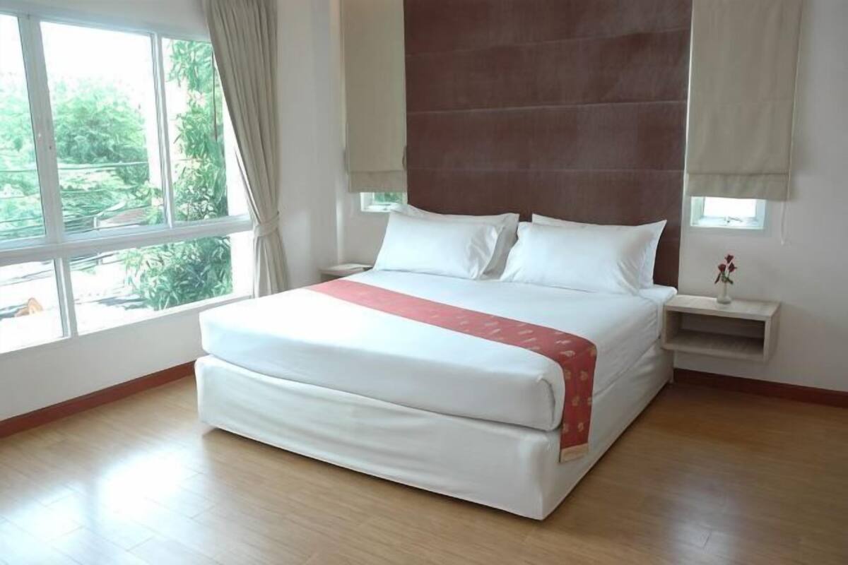 A large bed with a red blanket, a nightstand, a window with curtains, and a view of greenery outside.