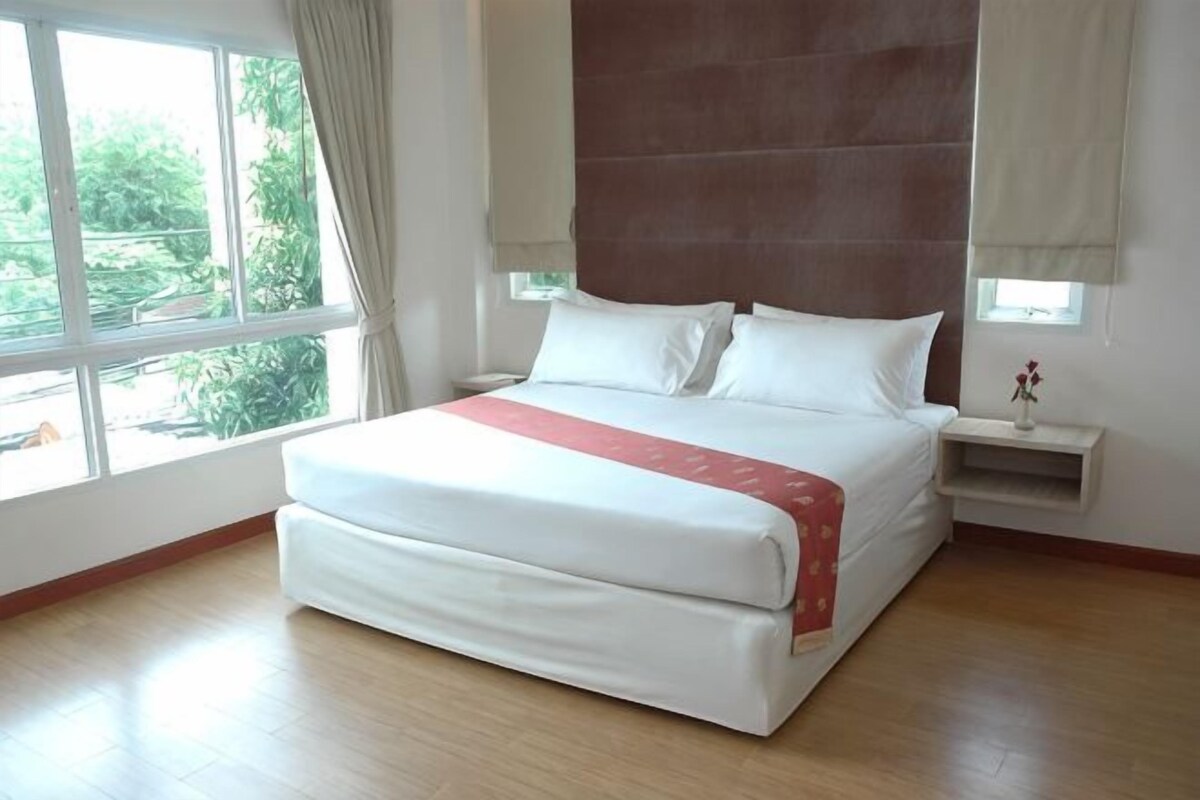 A large bed with a red blanket, a nightstand, a window with curtains, and a view of greenery outside.