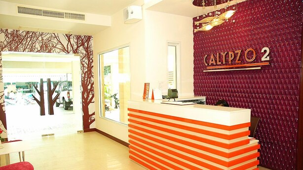 A reception desk with a red and white striped design, a sign reading "CALYPZO 2", and a red textured wall in the background. - Calypzo 2 (Bangkok)