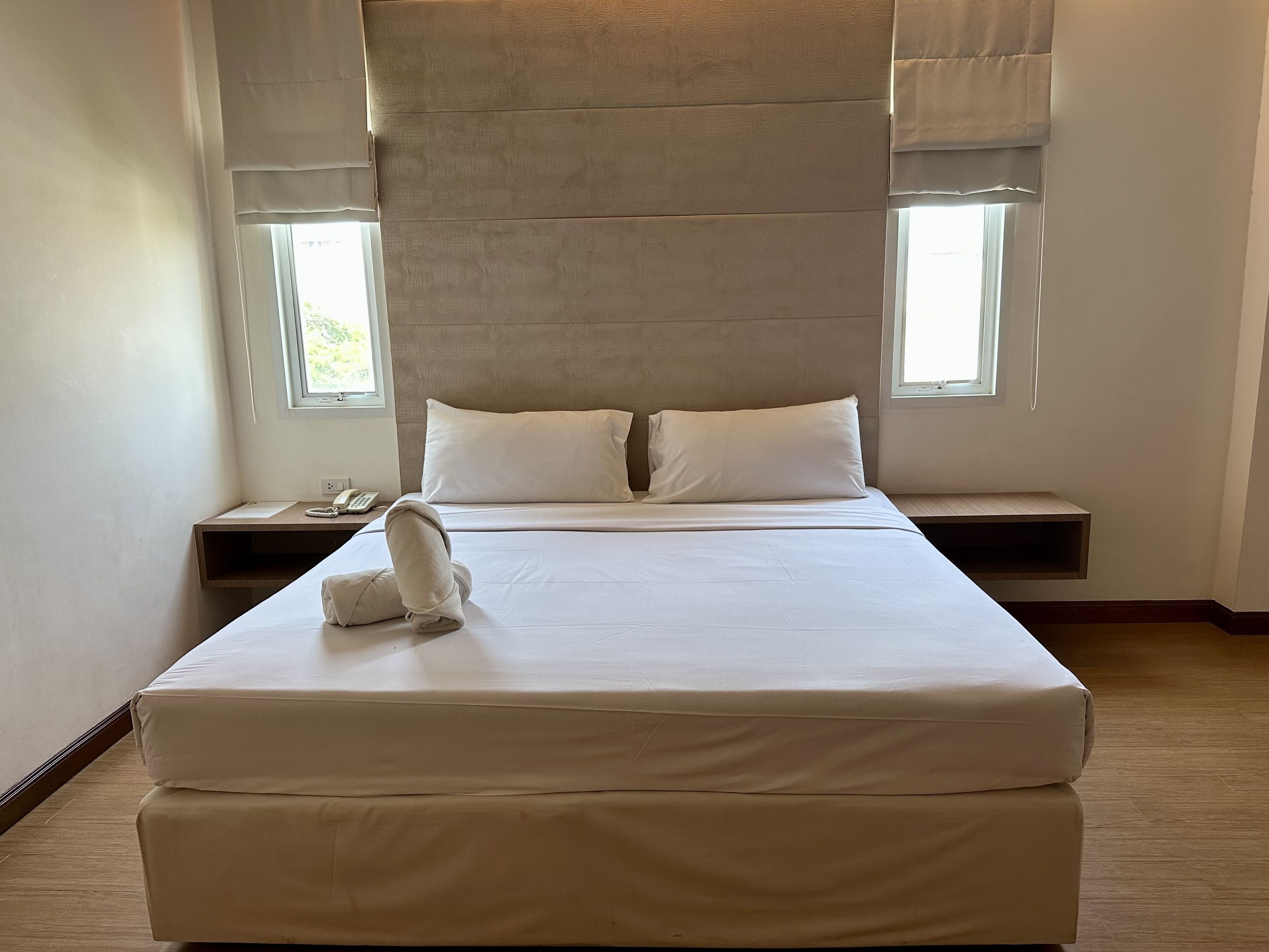 A neatly made bed with a pillow and a folded towel, a bedside table with a phone and papers, and a window with curtains.
