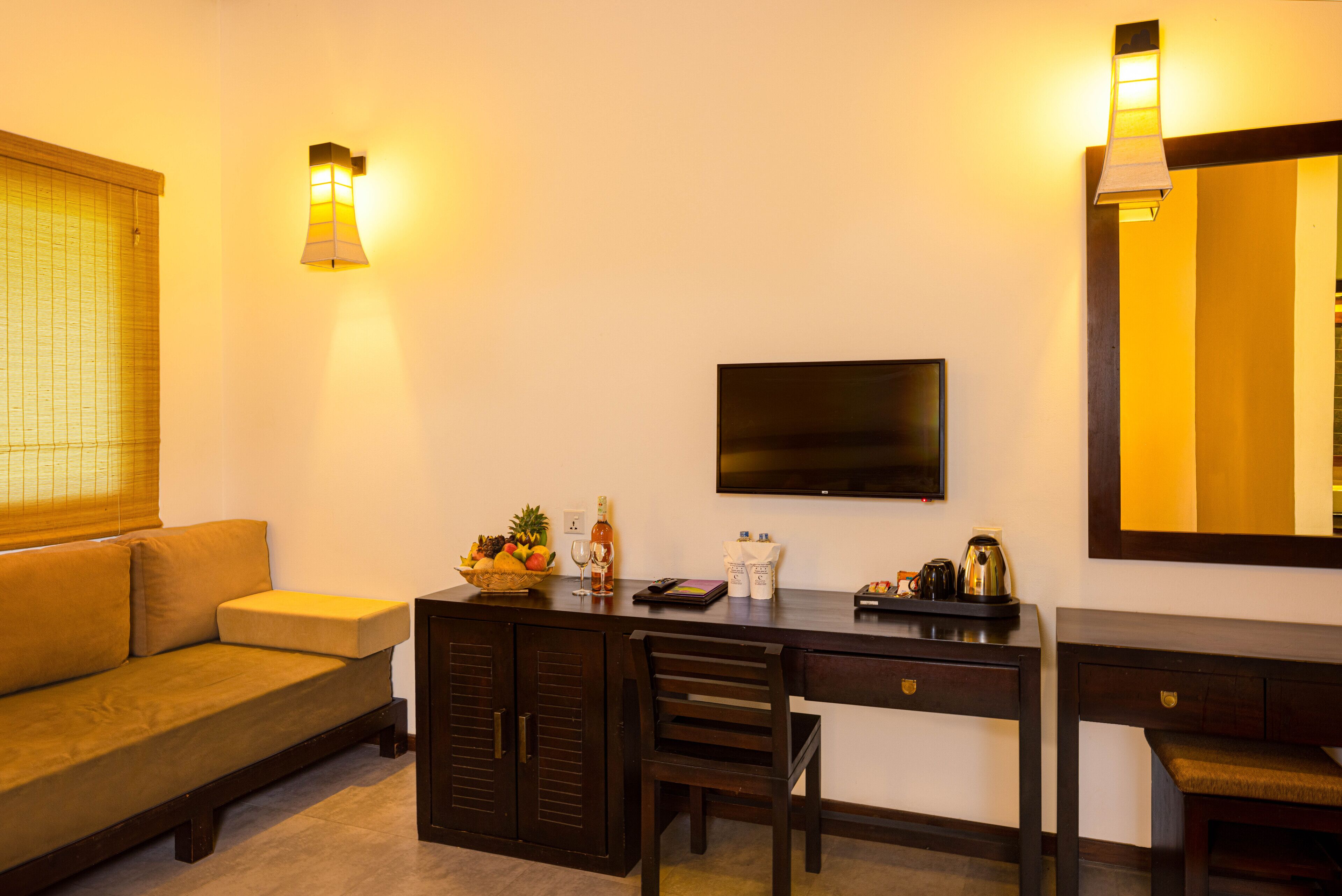 A hotel room with a sofa, a desk with a flat-screen TV, a mirror, and a small kitchenette area with a coffee maker and fruit bowl.