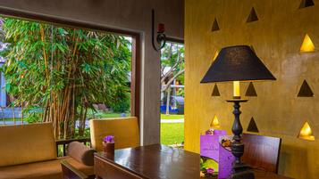 A dining area with a wooden table, chairs, and a lamp. There is a window with a view of greenery outside.