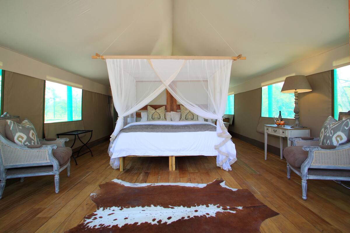 A spacious bedroom with a four-poster bed, two wicker armchairs, a small table, and a large cowhide rug.