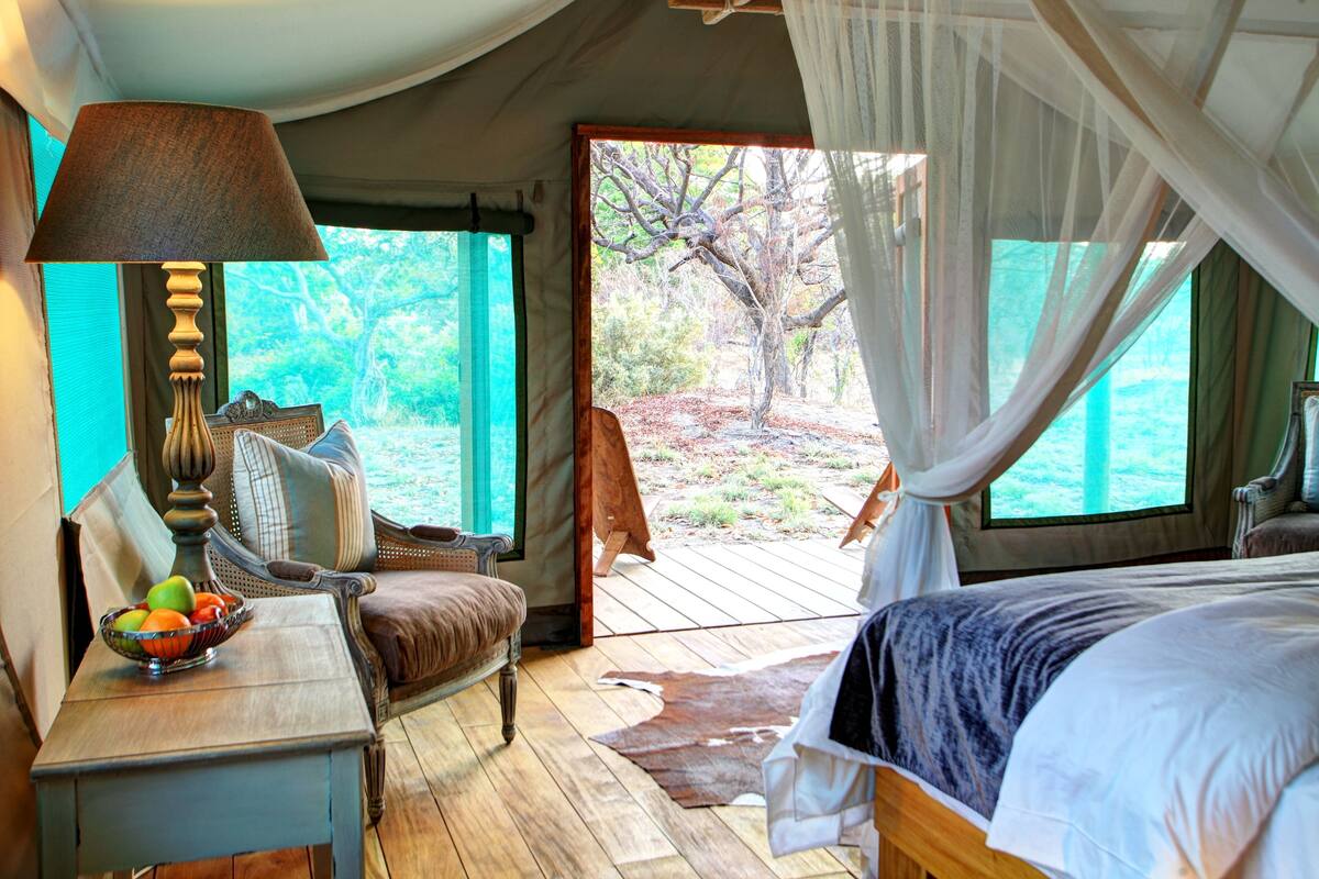 A tented room with a bed, a table with a lamp, and a view of the outdoors.