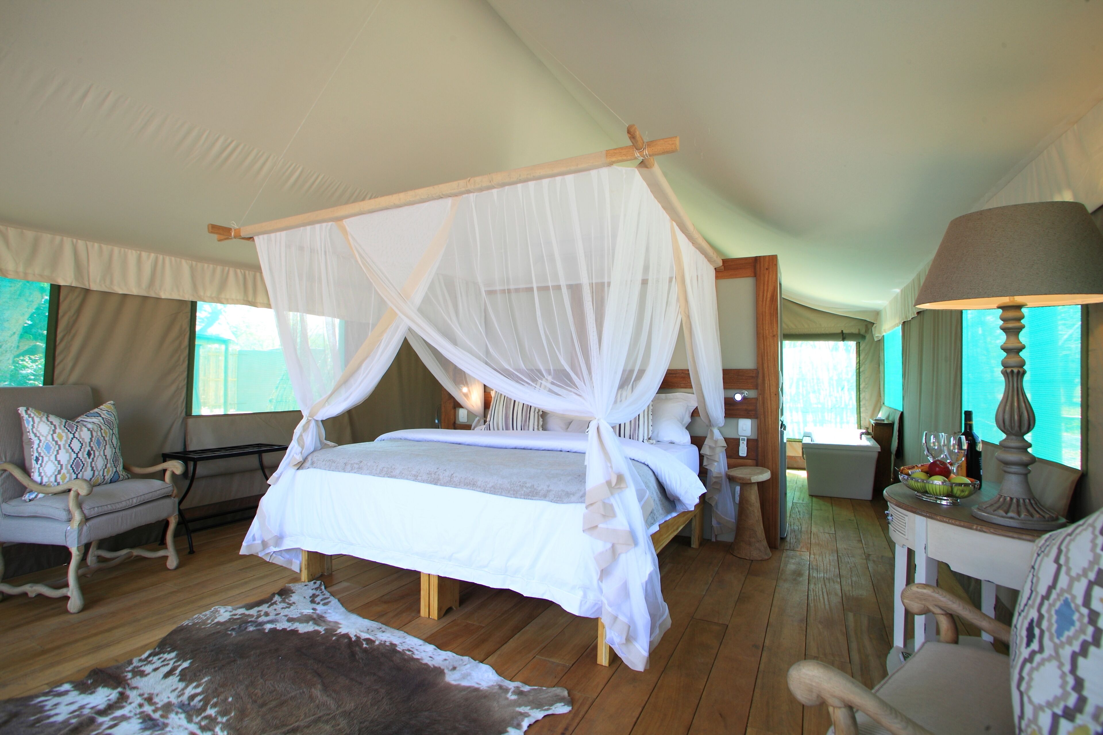 Un dormitorio amplio con cama con dosel, suelo de madera, alfombra de cuero de vaca, una silla, una mesa con lámpara y vistas al exterior.