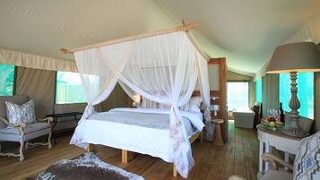 Un dormitorio amplio con cama con dosel, suelo de madera, alfombra de cuero de vaca, una silla, una mesa con lámpara y vistas al exterior.