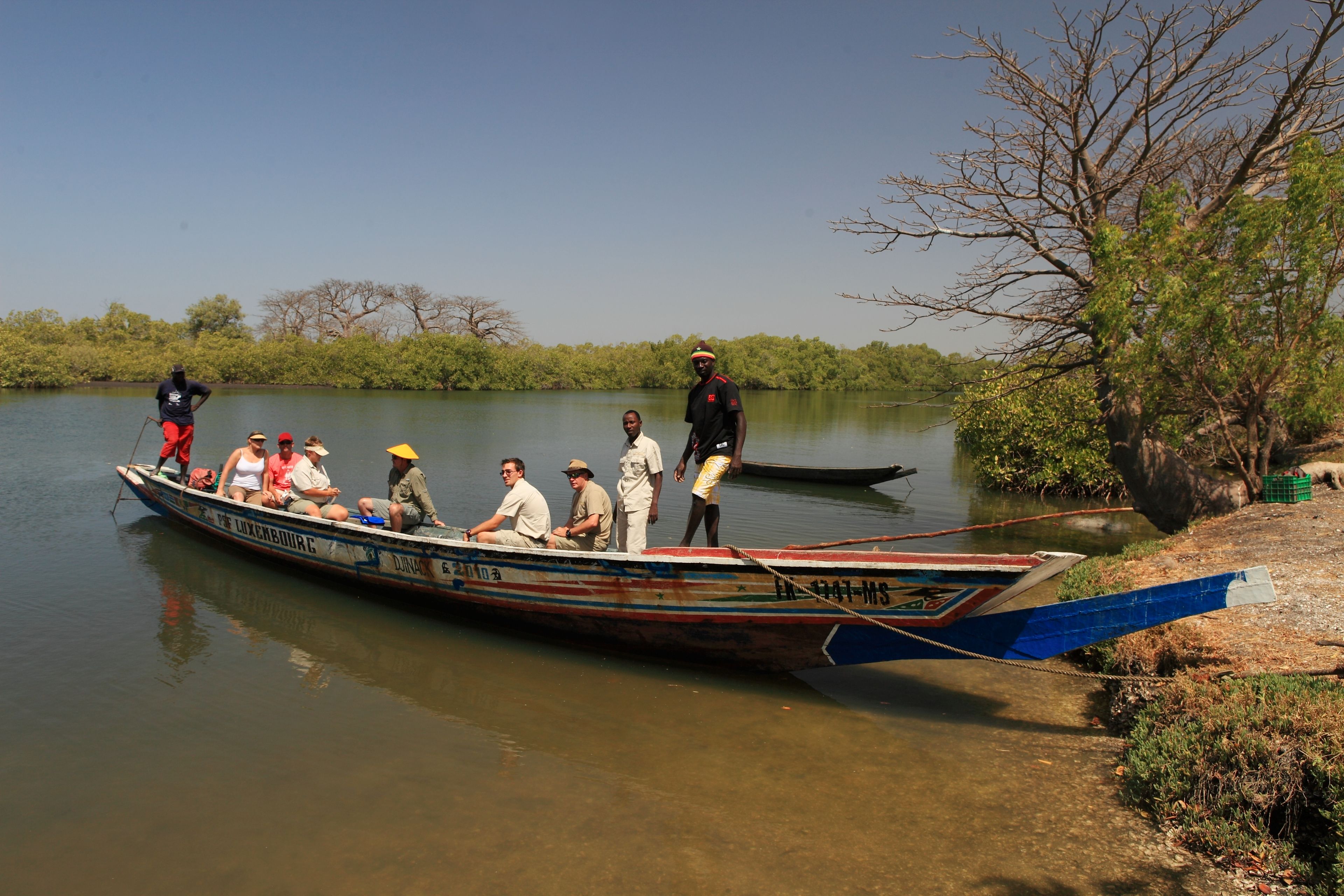 A boat with passengers and a guide, docked at a calm river with trees and greenery in the background.