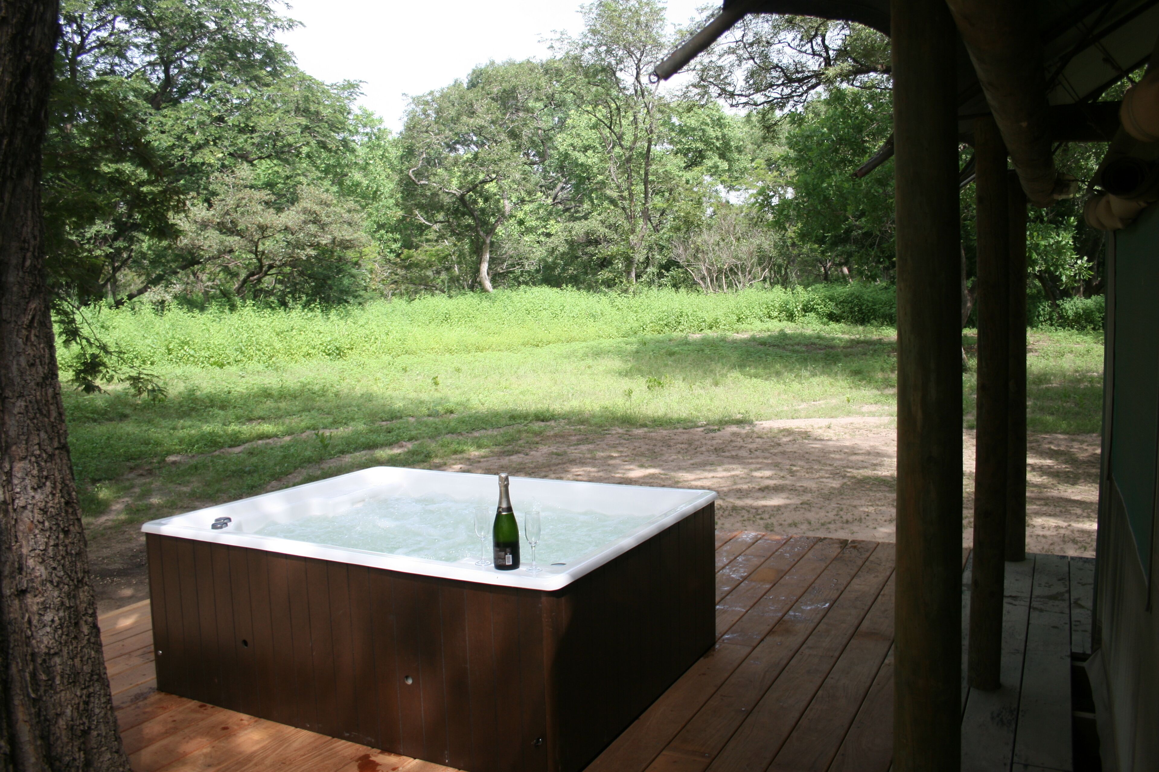 A hot tub on a wooden deck with a bottle and glass, surrounded by greenery.