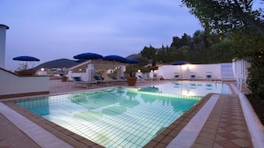 Piscina con sillas de descanso y sombrillas, rodeada de un solárium con baldosas y vistas a colinas.