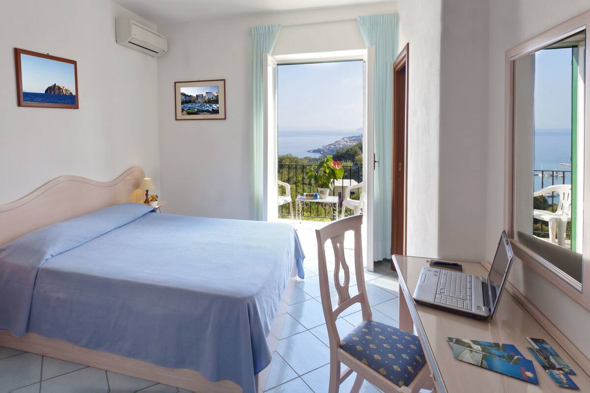 A bedroom with a bed, a desk with a laptop, a chair, and a balcony with a view of the sea.