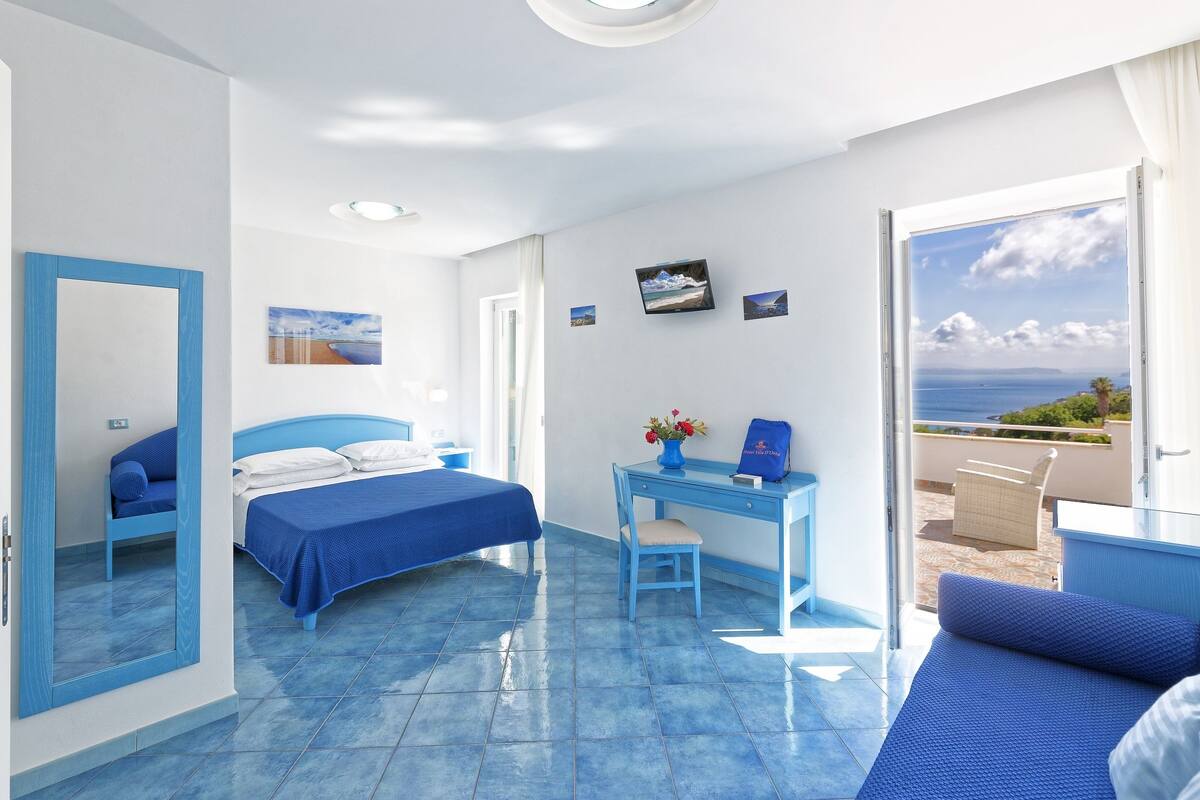 A room with a bed, a desk, and a chair, a large mirror, and a view of the sea.
