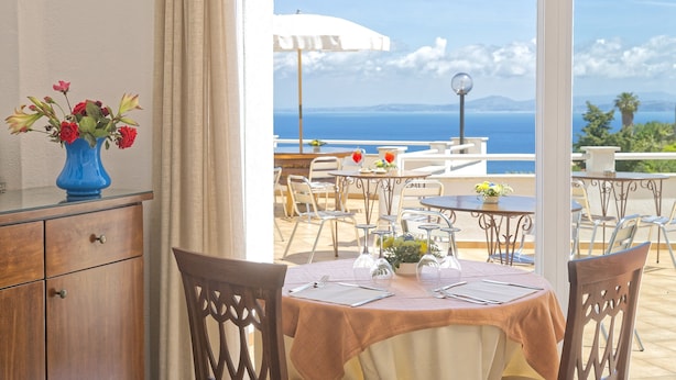 Un área de comedor al aire libre con mesas y sillas, un jarrón con flores y vista al mar.
