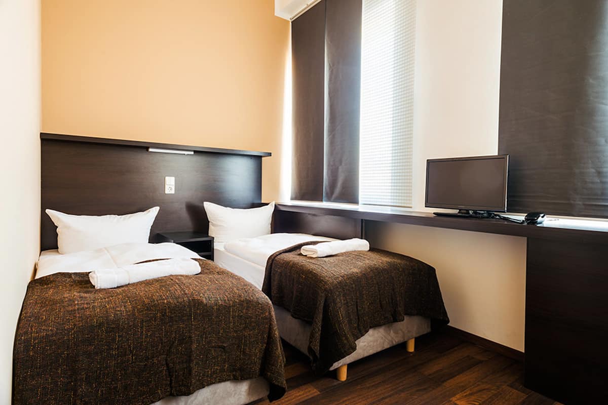 A hotel room with two single beds, a television on a stand, and a window with blinds.