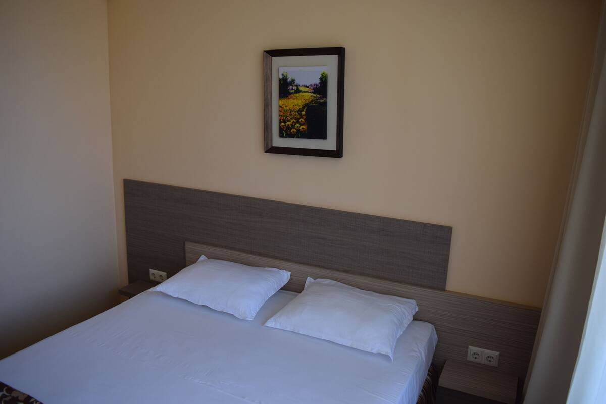 A hotel room with a bed, two pillows, a headboard, a nightstand, and a framed picture on the wall.