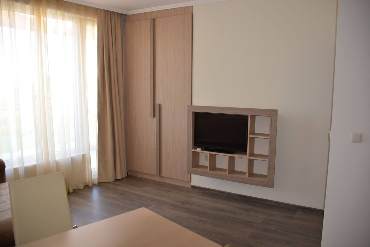 A room with a TV mounted on the wall, wooden flooring, a large window with sheer curtains, and a wardrobe.