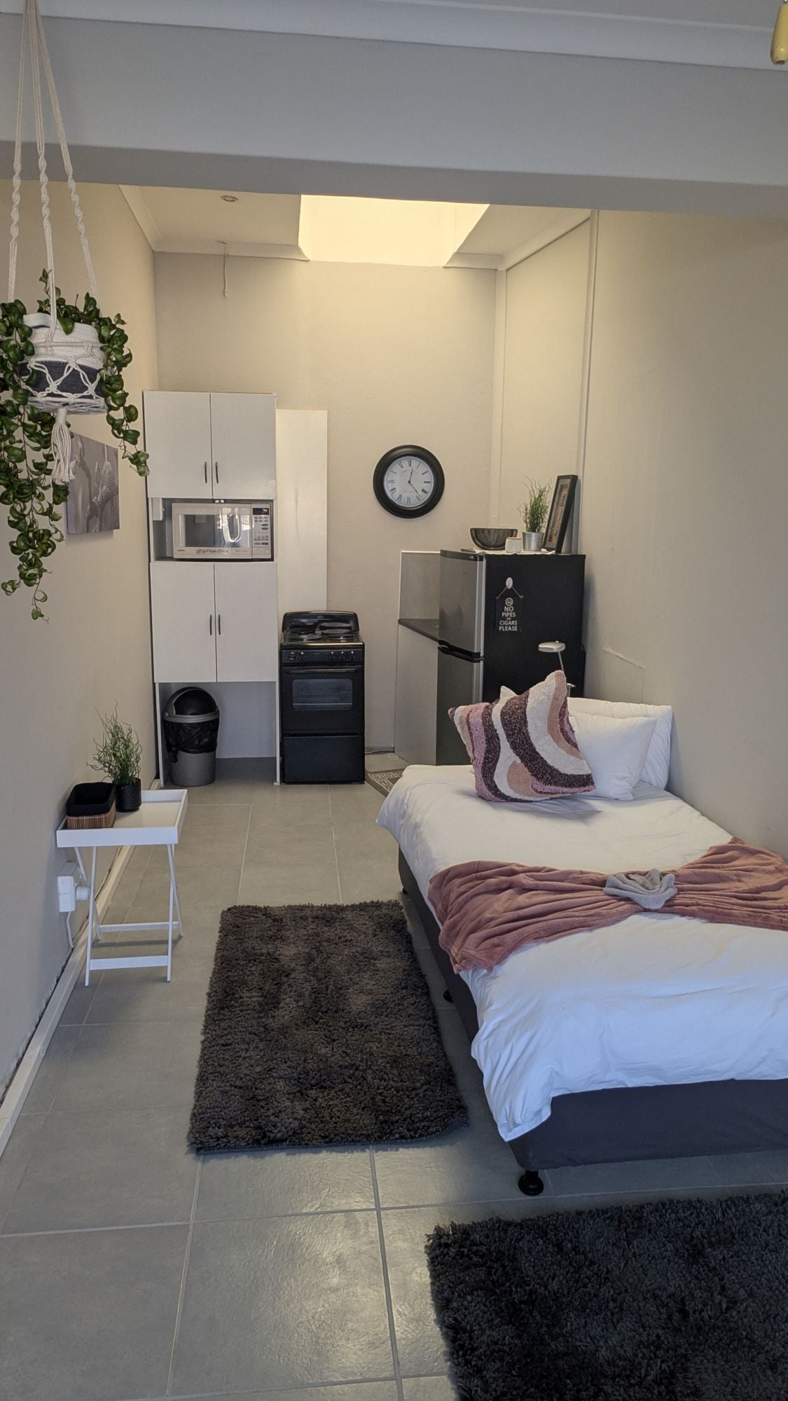 A modern bedroom with a bed, a small bedside table, a dark rug, a white wall, a clock, a microwave, a refrigerator, and a cabinet.