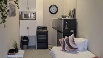 A modern bedroom with a bed, a small bedside table, a dark rug, a white wall, a clock, a microwave, a refrigerator, and a cabinet.