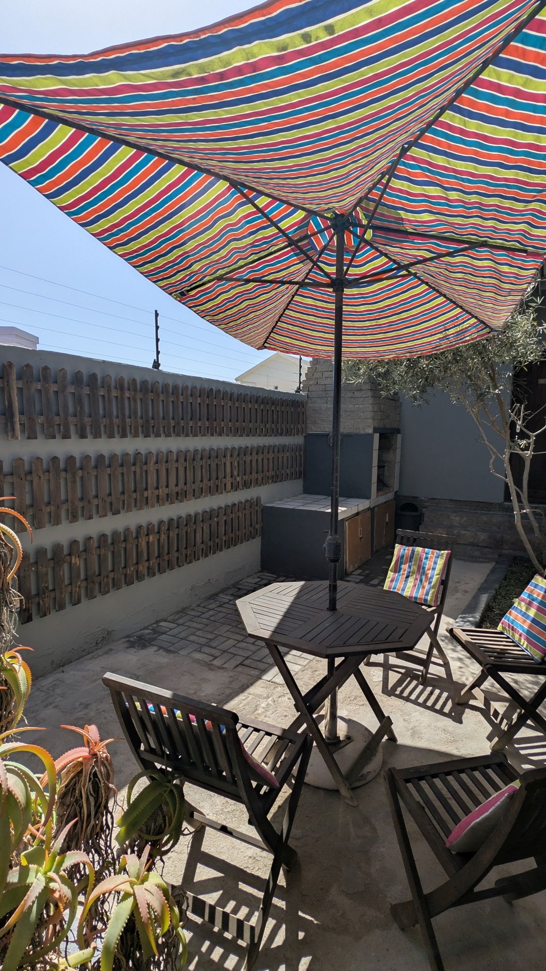 Een terras buiten met een streepjesparaplu, een barbecue en houten lounge fauteuils.