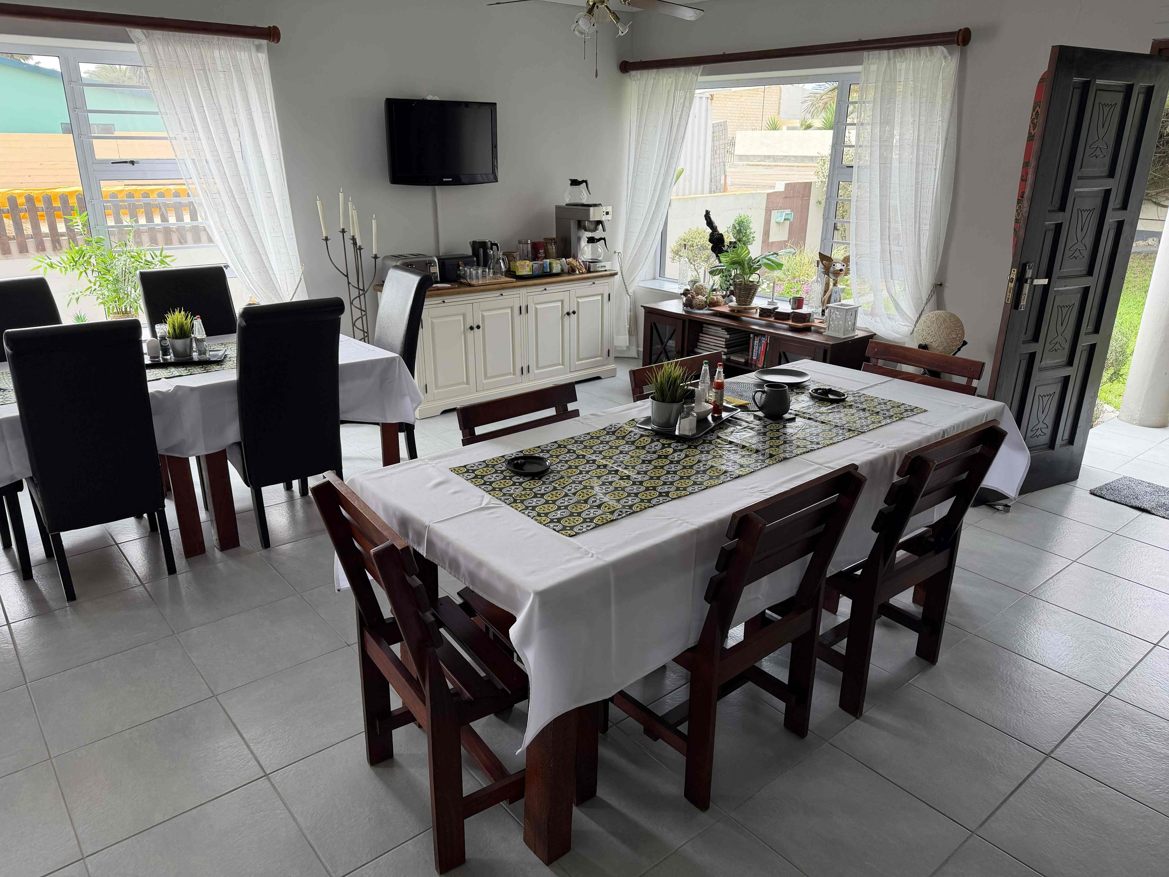 Un coin repas avec une table dressée pour six personnes, entourée de chaises, un téléviseur fixé au mur et une vue sur l’extérieur grâce à de grandes fenêtres.