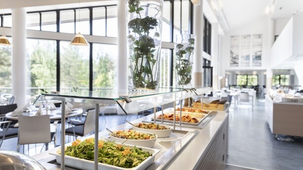 Zona de bufé con diversas opciones de comida, un expositor de vidrio y vistas al exterior a través de amplios ventanales.