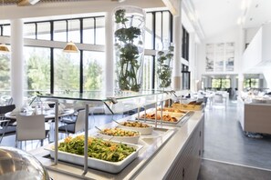 Zona de bufé con diversas opciones de comida, un expositor de vidrio y vistas al exterior a través de amplios ventanales.