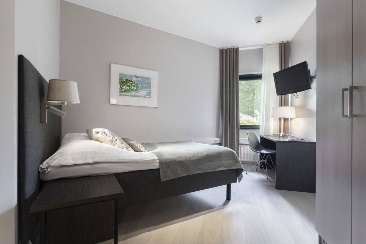 A modern bedroom with a bed, bedside table, desk, chair, and a flat-screen TV mounted on the wall.