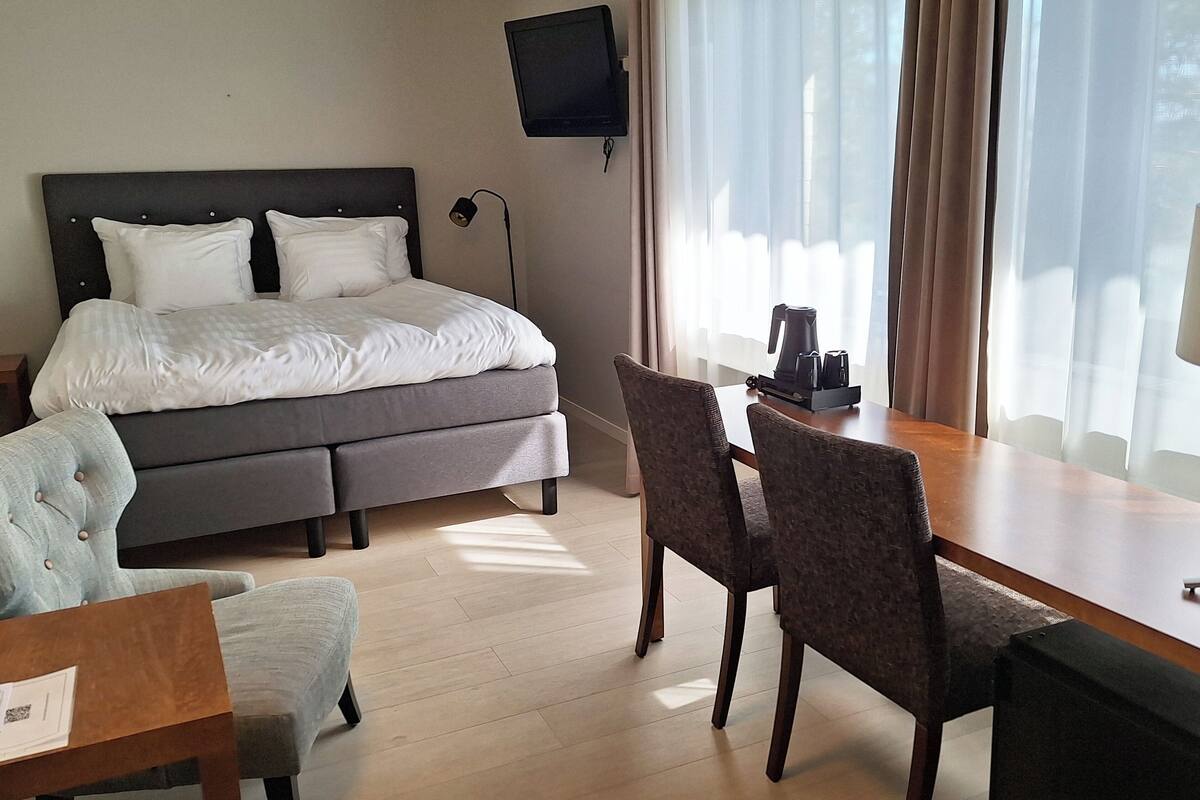 A hotel room with a bed, a desk with a chair, a TV mounted on the wall, and a window with curtains.