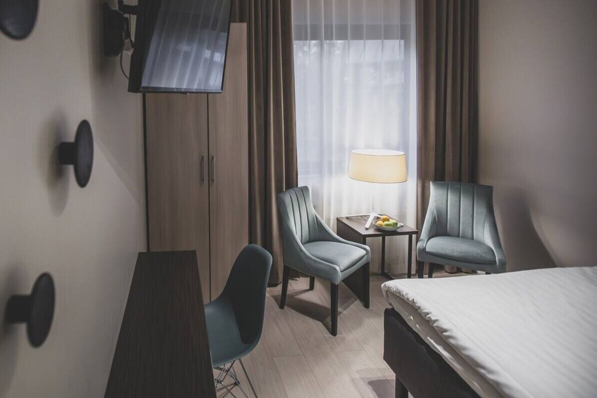 A hotel room with a bed, two armchairs, a small table with a lamp, a TV mounted on the wall, and a window with curtains.