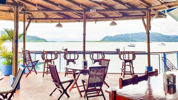 An outdoor seating area with wooden tables and chairs, overlooking a body of water with boats. - Fish Hook Marina & Lodge (Golfito)