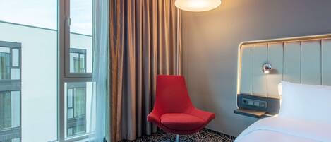 A hotel room with a bed, a red chair, a window with curtains, and a cityscape view.