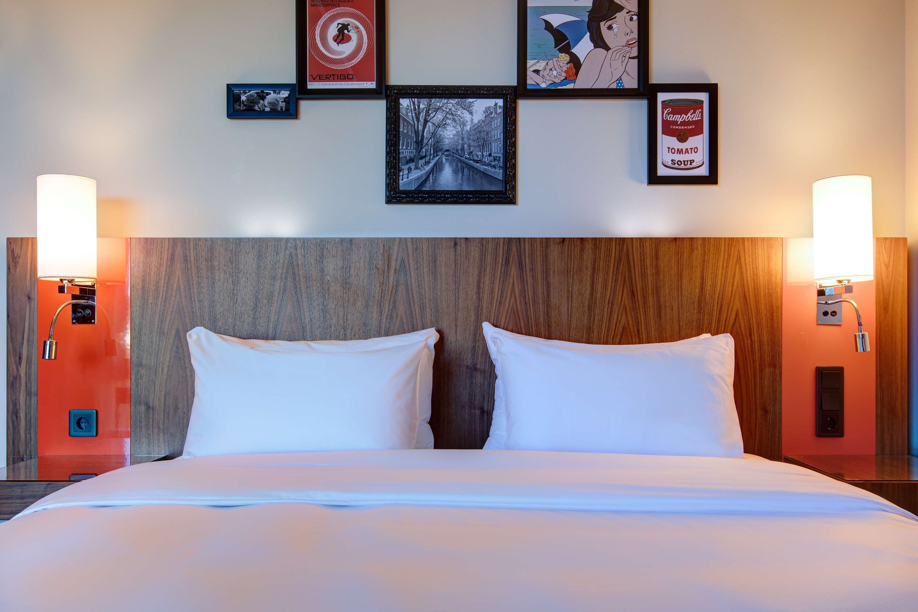 Habitación de hotel con cabecera de madera, dos almohadas blancas, cama con ropa de cama blanca y lámparas fijadas en la pared a ambos lados.