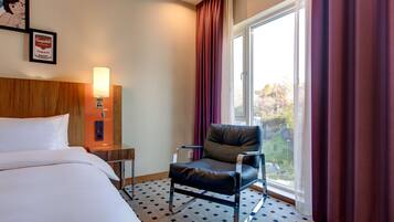 Une chambre d’hôtel avec un lit, une chaise, une table de chevet avec une lampe, un tableau encadré et une fenêtre avec des rideaux.