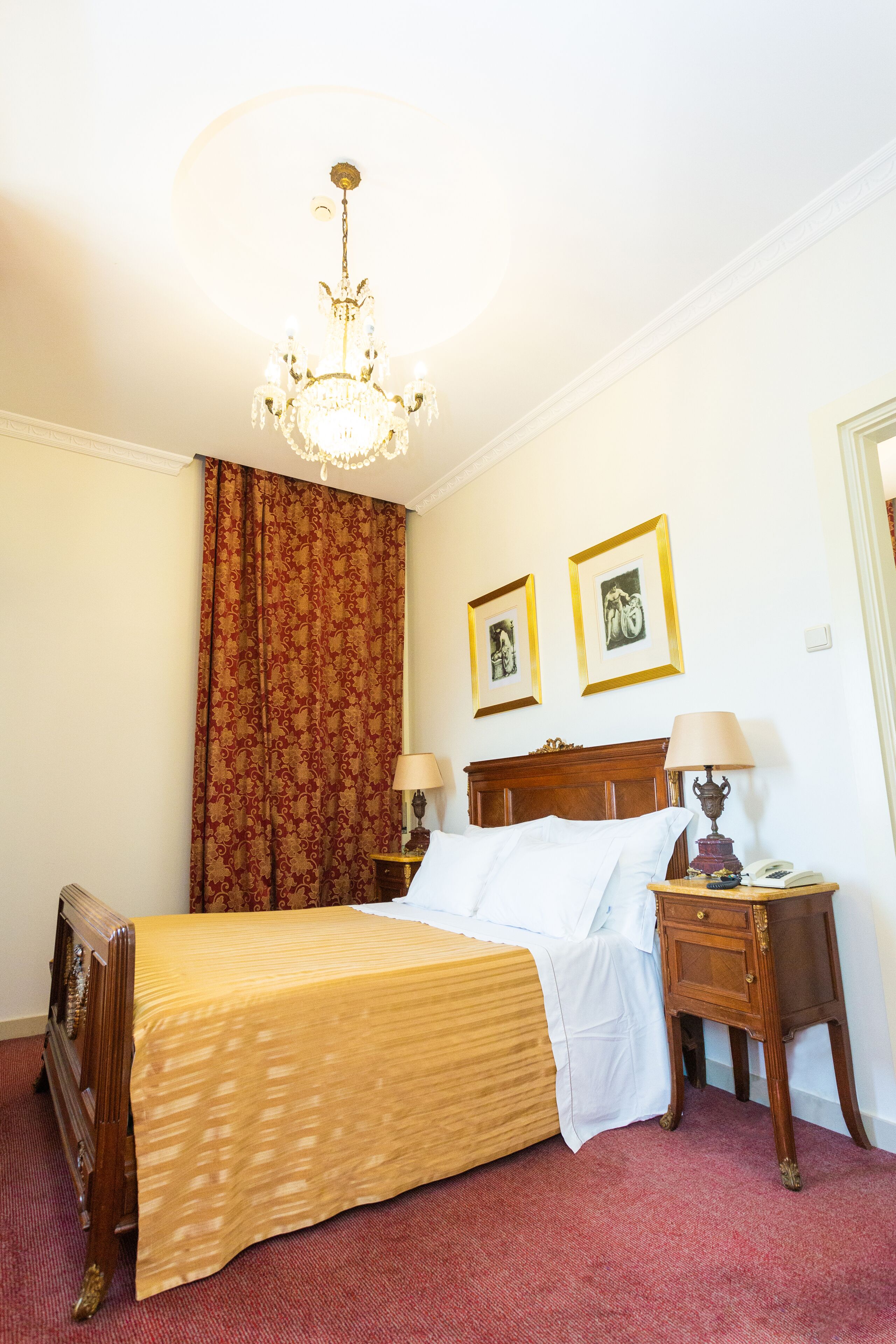 Un cuarto de hotel con una cama, una mesita de noche, una lámpara, una araña y dos cuadros en la pared.