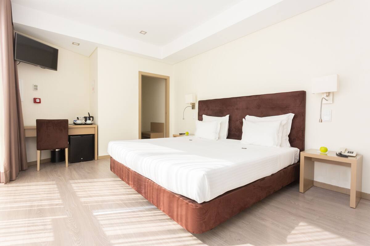 A hotel room with a large bed, a desk, a chair, a TV, and a bedside table with a phone and a fruit.