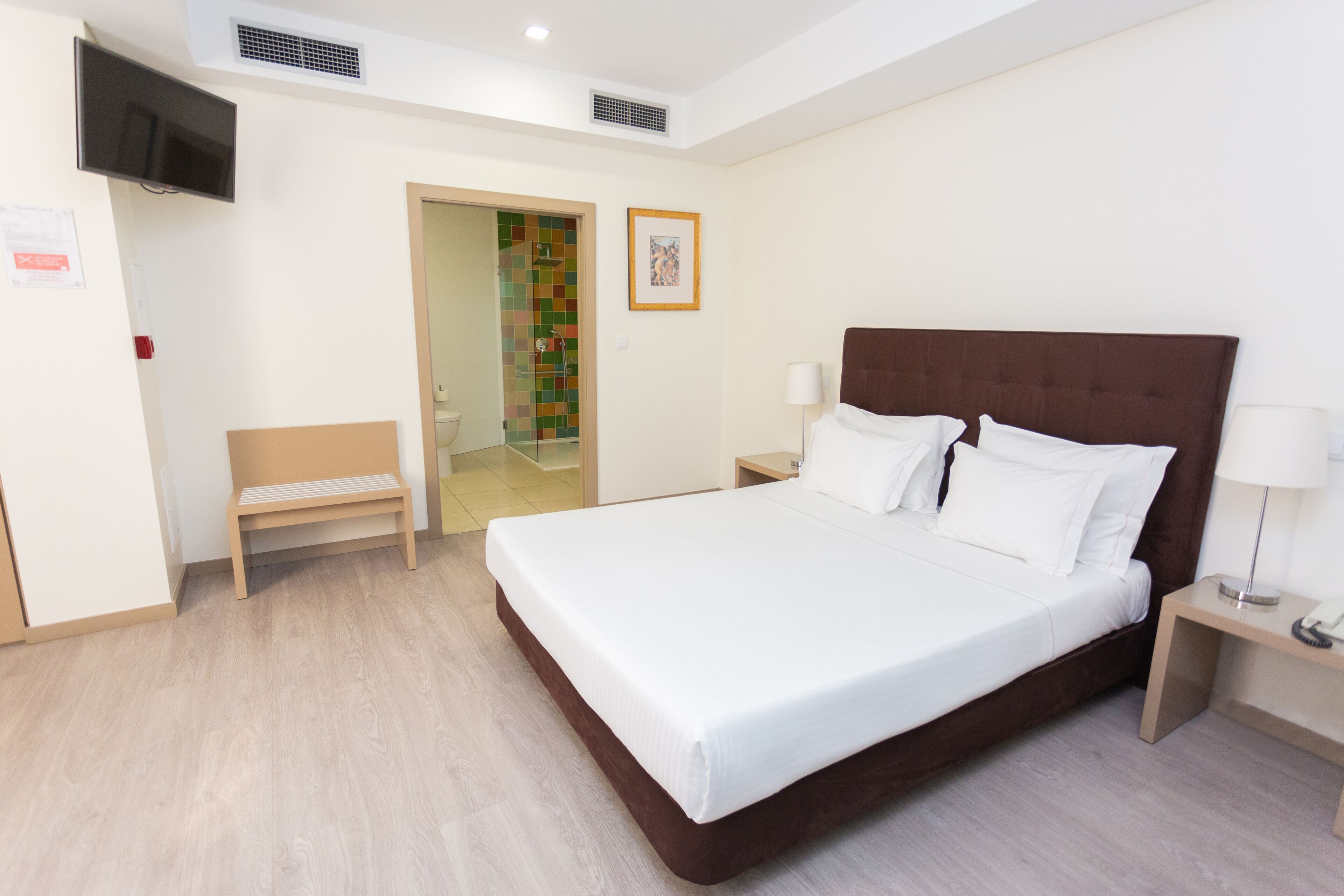 Habitación de hotel con una cama grande, mesitas de noche, un banco, un televisor y zona de ducha.