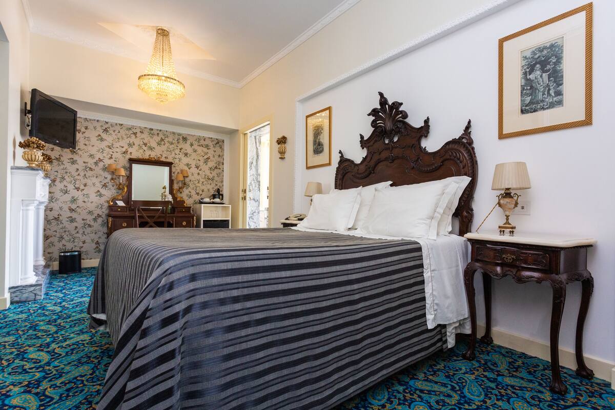 A hotel room with a large bed, a wooden headboard, a bedside table with a lamp, a dresser, a mirror, and a chandelier.