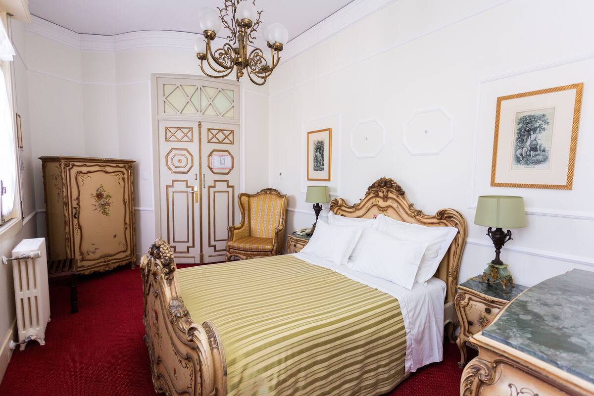 A classic room with a bed, a wooden cabinet, a chair, and a chandelier.