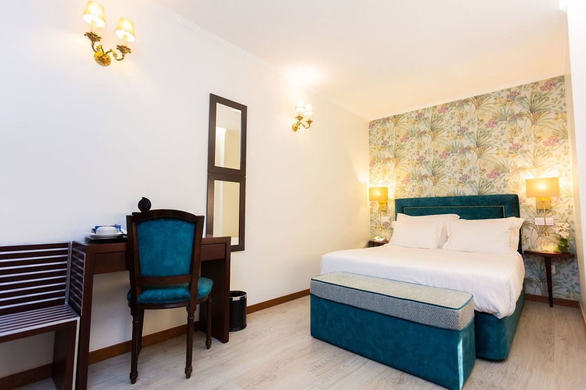 A hotel room with a bed, a desk, a chair, a trash bin, and two wall-mounted lamps.