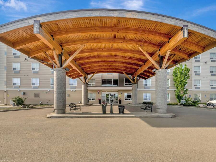 Un espacio exterior techado con techo de madera curva sostenido por columnas de concreto, frente a un edificio de varios pisos con fachada color beige y ventanas.