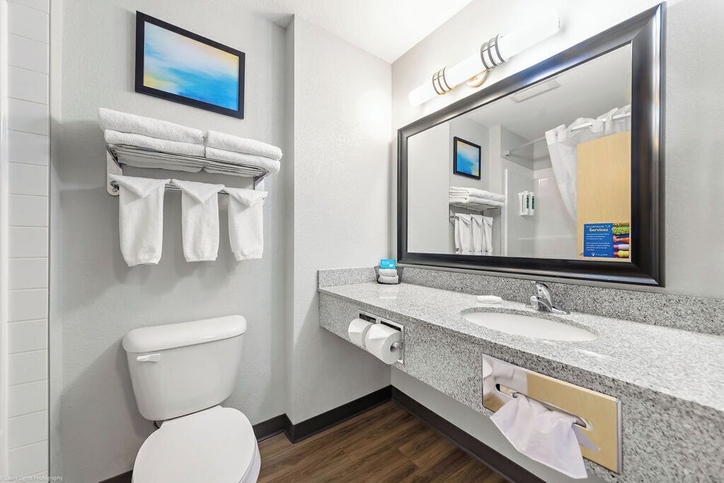 A modern bathroom with a toilet, sink, and large mirror. There are towels hanging on a rack and a framed picture on the wall.