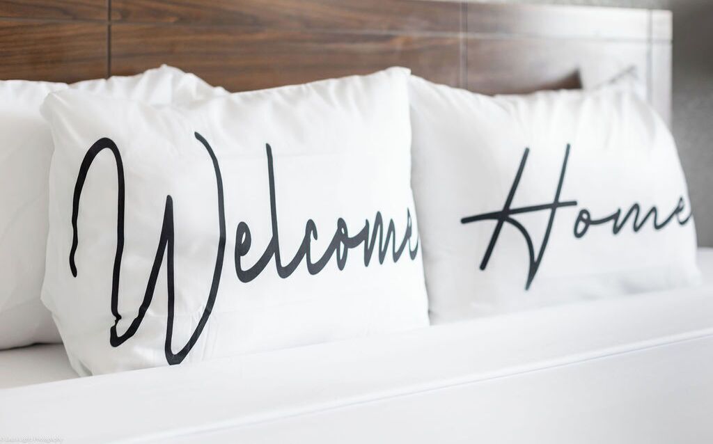 Una cama con dos almohadas, una con la inscripción 'Welcome Home' y la otra con la palabra 'Home'.