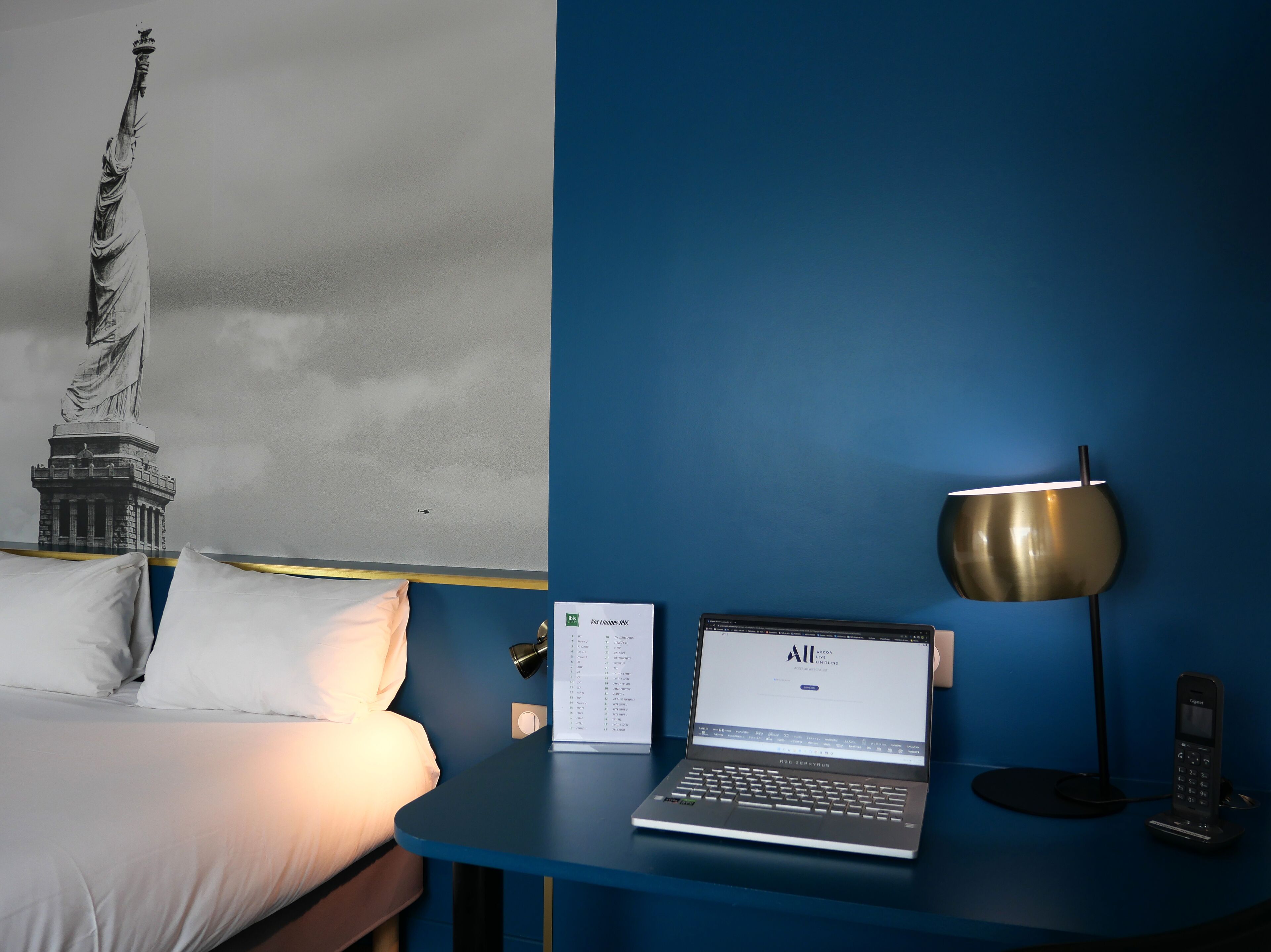 Une chambre d’hôtel avec un lit, un bureau avec un ordinateur portable, un téléphone et une lampe fixée au mur.