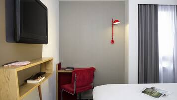 Una habitació d'hotel amb un televisor fixat a la paret, un escriptori amb una cadira, una cadira vermella, un llit i una finestra amb cortines.