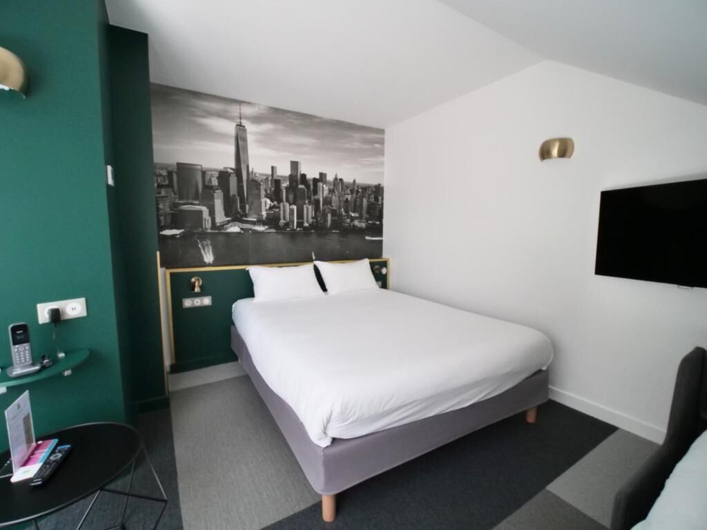 A hotel room with a bed, a desk with a phone and a tablet, a wall-mounted TV, and a cityscape mural.