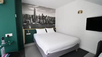 A hotel room with a bed, a desk with a phone and a tablet, a wall-mounted TV, and a cityscape mural.