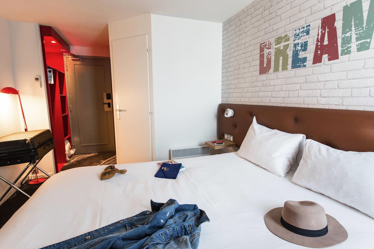 A hotel room with a white brick wall, a bed with white linens, a brown headboard, a nightstand with a lamp, and a red door.