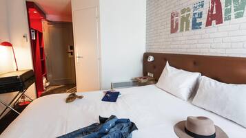 A hotel room with a white brick wall, a bed with white linens, a brown headboard, a nightstand with a lamp, and a red door.